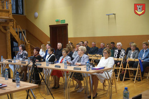 Siedzący przy stole Radni Rady Miejskiej, za nimi znajdują się siedzący goście - sołtysi, dyrektorzy, pracownicy urzędu (photo)