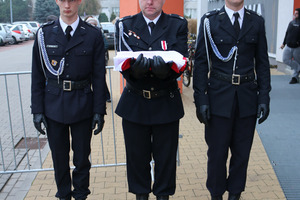 Trzej strażacy w oficjalnych mundurach. Mężczyzna stojący w środku trzyma w dłoniach flagę Polski (photo)